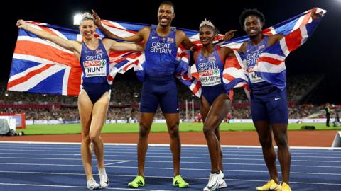 four people in running vests and shorts holding Union Jack flag standing on a track. All are smiling and looking directly at the camera. The women are wearing tiaras.