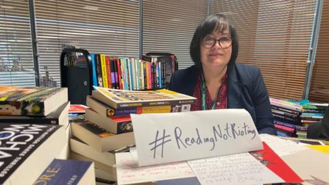 Denise Jones from Liverpool library service surrounded by books. She has short dark hair and is wearing glasses. A sign in front of her reads #ReadingnotRioting