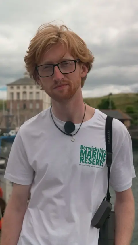 Patrick Safford at Eyemouth harbour, he has red hair, wears glasses and a white t-shirt with Berwickshire Marine Reserve on it