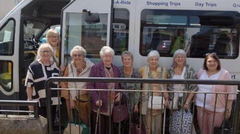 A group of older women dressed in casual clothes and smiling. They are standing next to a  small white bus.