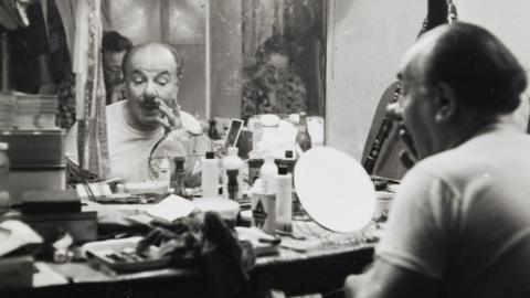 A black an white picture of Charlie Cairoli applying his face paint and nose. He is sitting at a cluttered dressing table in front of a large mirror.