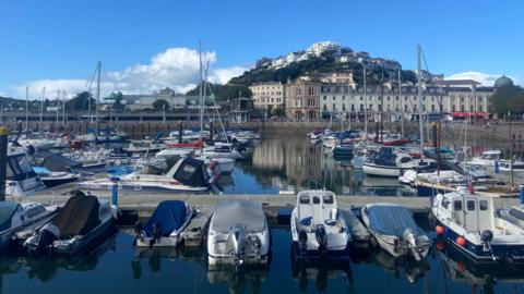 Dozens of boats moored in Torquay Harbour on a sunny day. Several buildings along the seafront of the town are in the distance.