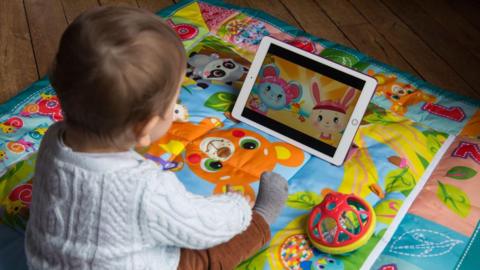 Toddler with its back to camera, on a playmat covered in animals and number in bright colours, watching a cartoon on an ipad. The ipad has two animated creatures.