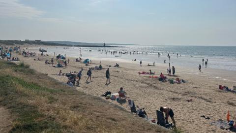 Beach with people on the sand and sea shore as well as swimming in the sea. The sun is shining and everyone is in swimwear or summer clothing. Some people have surf boards.