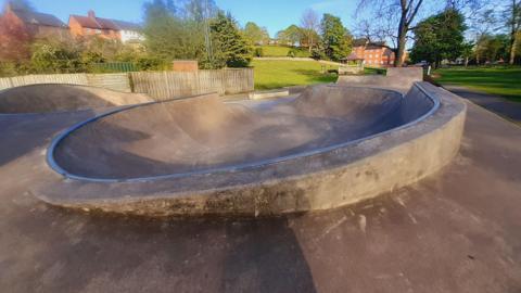 A contrate skate park in a park, surrounded by grass, trees, and houses overlooking the edge of the space.