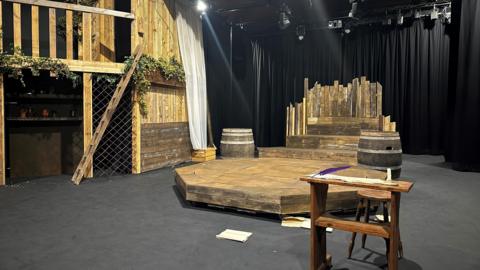 A wooden set on a stage. A platform is in the centre with two barrels and a fence on the other side. A chair and desk are positioned just in front of the platform. To the left is a wooden fence with plants halfway up.