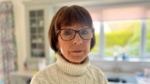 A woman with shoulder length brown hair looks at the camera, wearing brown rimmed, clear glasses. She wears a cream turtleneck. Behind her is a out of focus kitchen sink, with a window above it. A green hedge can be seen outside.