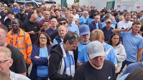 Large crowd of about a 100 people waiting. Most are wearing blue tops, including the City shirt. Many are looking frustrated or unimpressed, with some on their phones. A club steward in an orange stands to the left.