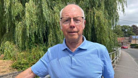 Councillor Paul McKernan wears a light blue polo top and black framed glasses. He has short grey hair and is looking towards the camera. Behind him is the road which traffic usually backs up on but in this photo it is quiet. There is just one red car on the road. To the left of him is a large willow tree taking up the majority of the shot.
