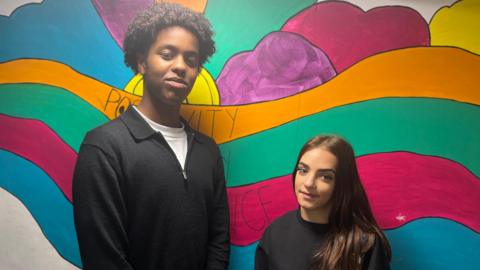 Ridwan and Jayda. Ridwan has short, dark curly hair. He is wearing a white t-shirt and a black jumper. Jayda has long dark hair. She is wearing a black jumper. They are stood side by side in front of a colourful wall. They are looking at the camera and smiling. 