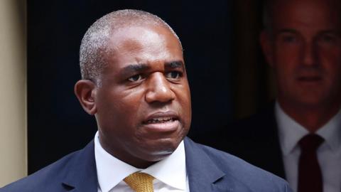 David Lammy wears a dark blue suit jacket, a white shirt and a yellow tie as he looks away from the camera in front of an open doorway