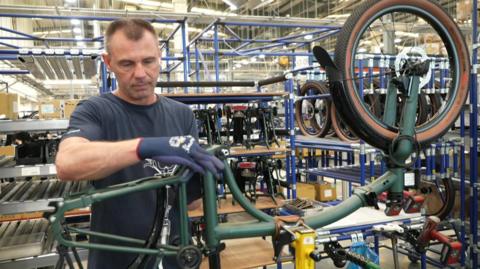A man is stood in Brompton bike factory. He has a navy blue t-shirt on and gloves and he is working on a dark green bike frame. 