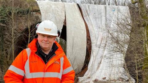 A man, Chris Bond, is standing in front of a damaged bridge. He is wearing a white hard hat with "Severn Valley Railway" written on it, and an orange high-visibility jacket with his name written on one side, with "Severn Valley Railway" on the other. The bridge behind him is covered in three huge white sheets. There are trees and shrubbery either side.