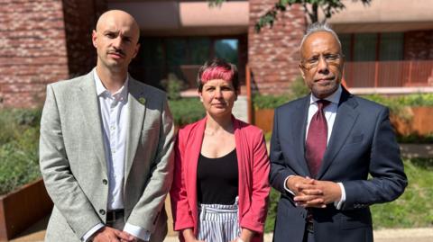 Three people stood in a line, from left man wearing light grey jacket, in the middle a woman wearing a light pink blazer, and a man wearing a navy suit. Behind them is parkland and tower blocks. It's a sunny and bright day. 