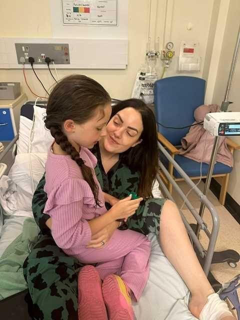 Lady on a hospital bed, dressed in green patterned pyjamas, smiling at her young daughter who is sitting on her lap. 