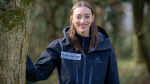 Camilla Hempleman-Adams stood centre, smiling in the camera. She is leaning on the trunk of a tree.