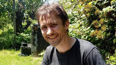 A picture of a man in a black T-shirt smiling in a sunny garden. He has dark brown hair with a fringe and a stubbly moustache and beard.