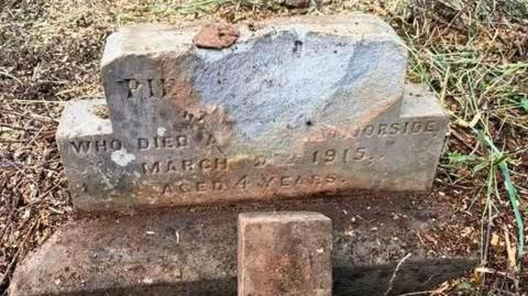 Two small, weathered gravestones stand in grass and earth. The upper stone is broken at the top, with faint, eroded lettering noting a death in 1915 at age four. Below it sits a smaller cross‑shaped marker inscribed with “In Memory.”