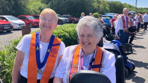 Two women enjoying the parade on a very sunny day in Rossknowlagh, County Donegal. The woman on the left has short blonde hair and glasses and the woman on the right has short, grey hair. They are both wearing short-sleeved white tops, orange collarettes and they are smiling broadly.