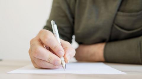 A male hand holding a pen, writing on paper. The upper body is visible (but not the face) and is wearing a green jumper.