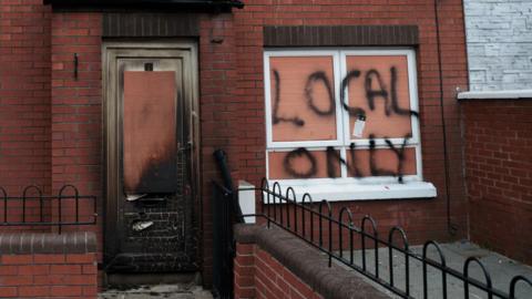 A view of the vandalised house.  The red-brick terraced property has windows on either side of the front door on both its ground and upper floors.  The downstairs windows have been boarded up and the words "local only" have been sprayed on the right hand side window.  The white PVC front door has sustained smoke and fire damage. 