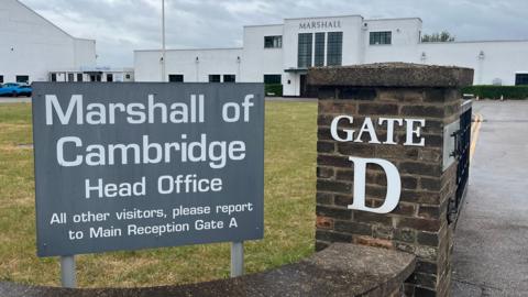 A large grey sign in the foreground says "Marshall of Cambridge Head Office". It is next to a brick pillar with "Gate D" on it. In the background is the company's white Art Deco reception building.
