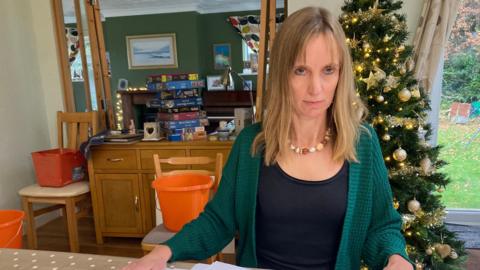 Hayley Jones sitting at her kitchen table with a pile of paperwork from the case in front of her. Behind there are buckets in the kitchen to catch water leaking from the ceiling above.