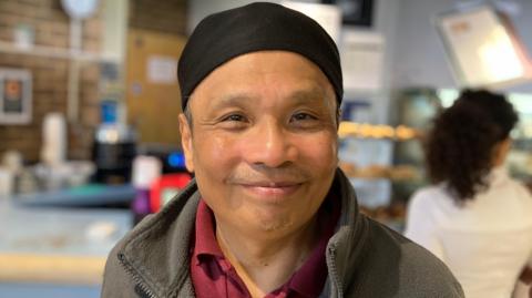 A man in a grey jacket and black hat smiles at the camera with the cafe blurred in the background. 