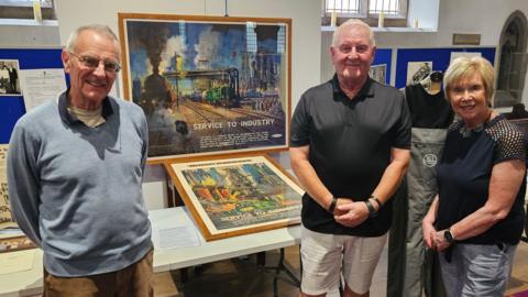 Three people; to the left is an elderly man with receding grey hair. He is wearing a pale blue jumper or polo shirt over a white t-shirt. To his right is an elderly man, also with receding grey hair and wearing a short-sleeved black shirt and white shorts. On the far right is a woman with short blonde hair, wearing a black top and light blue shorts or trousers. All three are smiling. Behind them are stands with old photographs and documents, and in a prominent position between them are two colourful old railway posters.