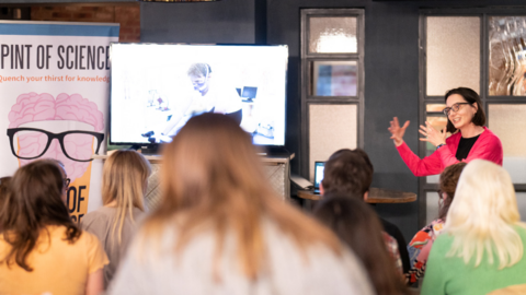 A woman is addressing a seated crowd and giving a public lecture, she is explaining and using her hands to gesture to illustrate her talk. Next to her,
there is a large screen with images in it.