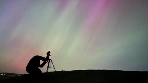 A man stands with a tripod and a camera pointing it up to the sky which is green and red with the northern lights.