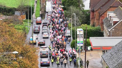 A drone shot of a protest held in Crowborough, East Sussex on 16 November over plans to house asylum seekers at an army site in the town. Hundreds of people can be seen walking on the streets with flags and banners. Cars are driving alongside the walkers. 