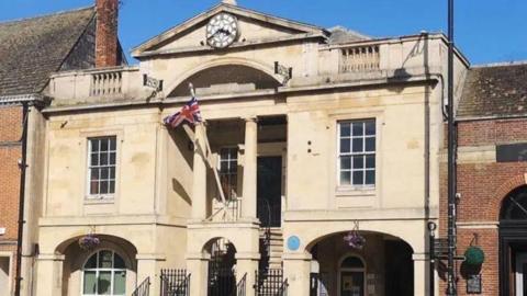 An outside view of the old town hall. It is a beige tall building with four plinths which create a dramatic entrance. A union jack flag which is attached to an archway flutters in the wind.
