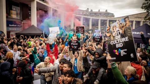 Hundreds of people gathered in Bradford City Park. They are holding up signs that read 'UK City of Culture Bid' and cheering as they celebrate the news that Bradford will be UK City of Culture in 2025.