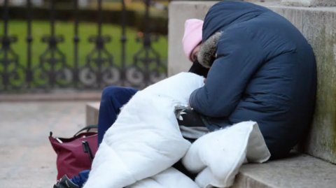 Two people are rough sleeping on some steps. One is wearing a blue jacket, the other a pink hat. 