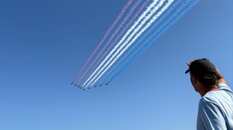 A man wearing a blue t-shirt and a black baseball cap shades his eyes as he watches the Red Arrows fly overhead, trailing red, white and blue smoke across a blue cloudless sky.
