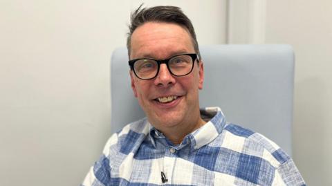 Paul Silvester smiling in a chair wearing a blue and white checked linen shirt and black thick-rimmed glasses
