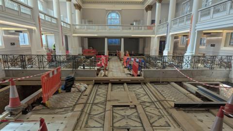 Interior of a neoclassical building with ornate columns and a mosaic tile floor undergoing renovation, with wooden planks, traffic cones, and barriers in place.