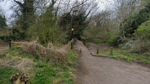 A country lane where the road comes to an end and becomes a footpath. There are trees and bushes surrounding the path