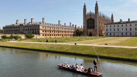 A view over the River Camb towards King's College Chapel and other ornate college buildings. A punt with around a dozen sightseers on board is seen in the foreground, being pushed along the river by a punting chauffeur, who is wearing a white T-shirt and waistcoat. He is holding a long metal staff, which is used to push against the bottom of the river and propel the boat along. Beyond the river are lawns leading up to the buildings in the middle distance. The sky is blue and it appears to be a gloriously sunny day.