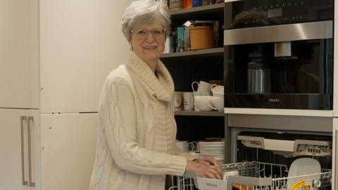 Judith Leary-Joyce unloading her dishwasher while smiling at the camera. She has white hair and glasses is wearing a long white knitted jumper.