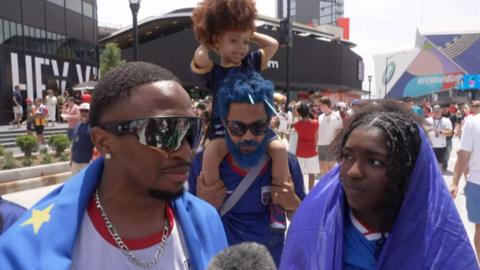 Four Cape Verde fans wearing blue and red, including a child sitting on a man's shoulders, stand to speak with a reporter.