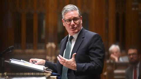 Keir Starmer speaking at the despatch box the House of Commons, he has a folder with notes and a microphone in front of him 