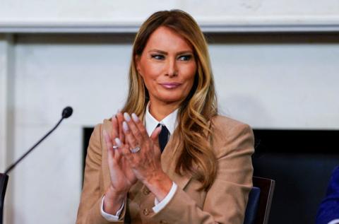 U.S. first lady Melania Trump wearing a tan suit sitting in front of a microphone and applauding. She is smiling and her hair falls over her shoulders as she looks off to one side