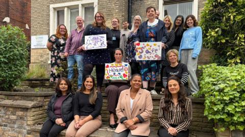 A group of people pose together outside a brick building, with some standing and others sitting on steps, holding colourful hand‑made signs with words and drawings.
