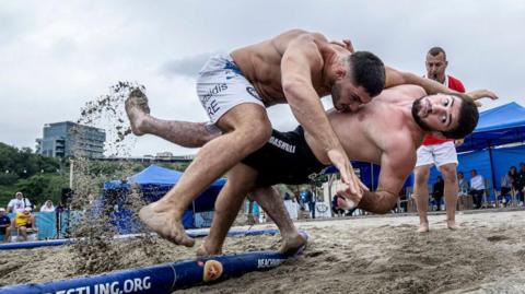 Two athletes compete in a beach wrestling match on a sandy outdoor ring. One athlete lifts and drives the other toward the edge of the arena, sending sand flying into the air. A referee stands nearby watching the action. Blue tents, spectators, and buildings are visible in the background under an overcast sky. 