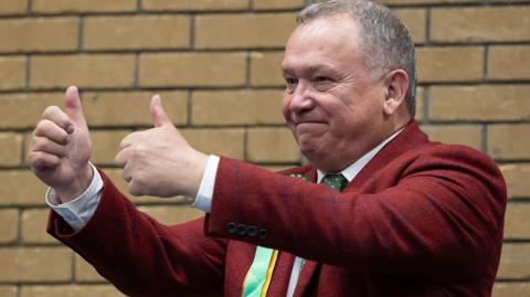 Plaid Cymru's newly elected Caerphilly Member of the Senedd Lindsay Whittle at the count. He is wearing a maroon suit jacket with blue check marks and a green tie with a white shirt. He is giving a double thumbs up and is smiling. Lindsay is a middle aged man with short grey hair