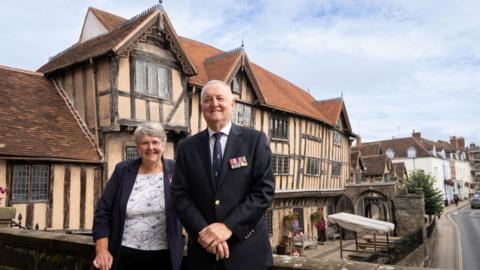 A man and a woman, both wearing navy jackets with military medals on them, stand in front of a Tudor building painted yellow with exposed wooden beams