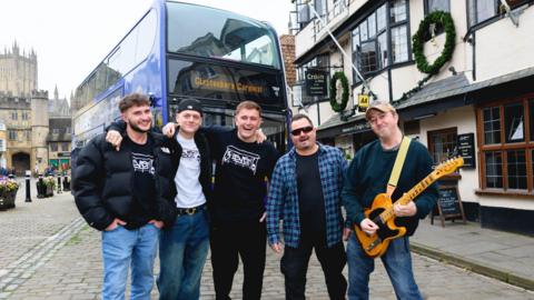 3SEVEN6 members and Bad Cowboys members are standing next to each other in front of a bus. They are by a pub in Wells. 