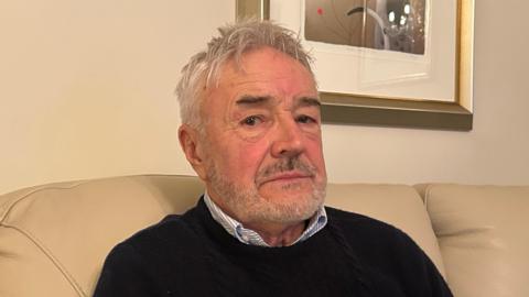 A close up picture Jim Rowe who is sat on a cream soft looking at the camera. He is wearing a black knitted jumper with a blue and white striped shirt underneath. Behind him there is a piece of artwork in a gold frame placed on a white wall.